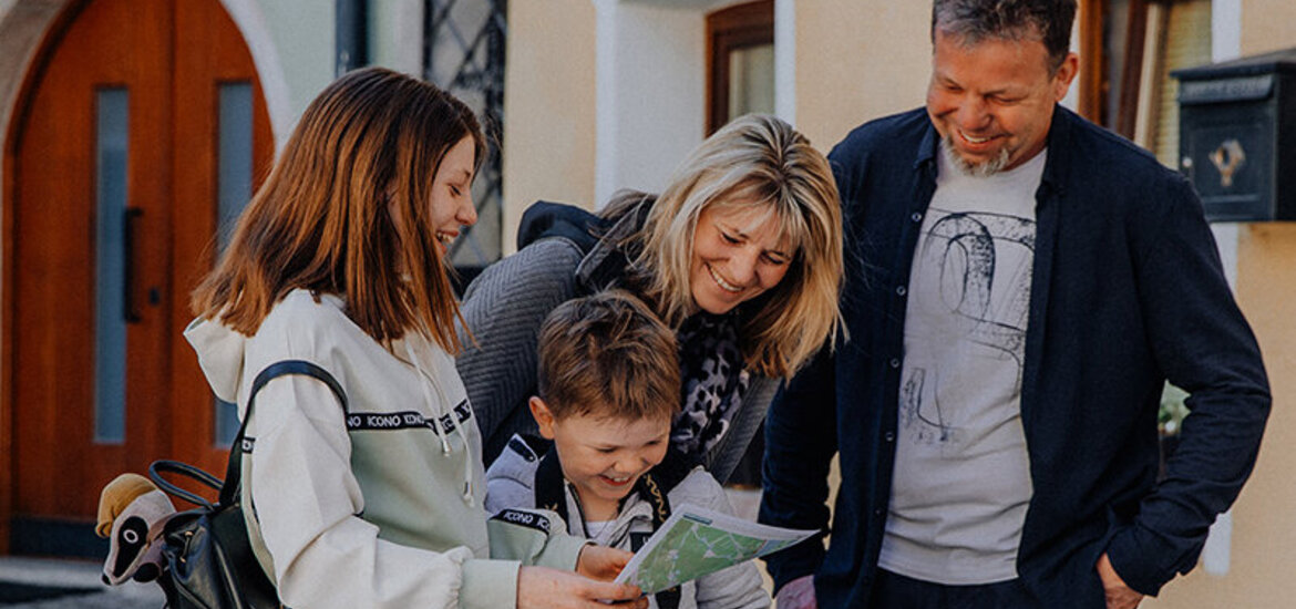 Familie mit zwei Kindern steht auf Kopfsteinpflaster und betrachtet gemeinsam Karten und Ausdrucke, der Junge hält eine Kamera in der Hand