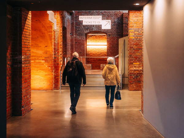 Zwei Personen gehen durch einen Gang mit roten Backsteinwänden im Dokumentationszentrum Reichsparteitagsgelände, darüber ein Schild mit der Aufschrift 'INTERIM TICKETS' und einem Pfeil nach rechts.