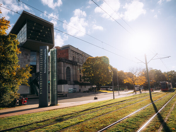 Das Dokumentationszentrum Reichsparteitagsgelände mit dem markanten Eingan links, im Hintergrund die Kongresshalle, rechts Straßenbahnschienen und herannahende Straßenbahn bei sonnigem Herbstwetter.