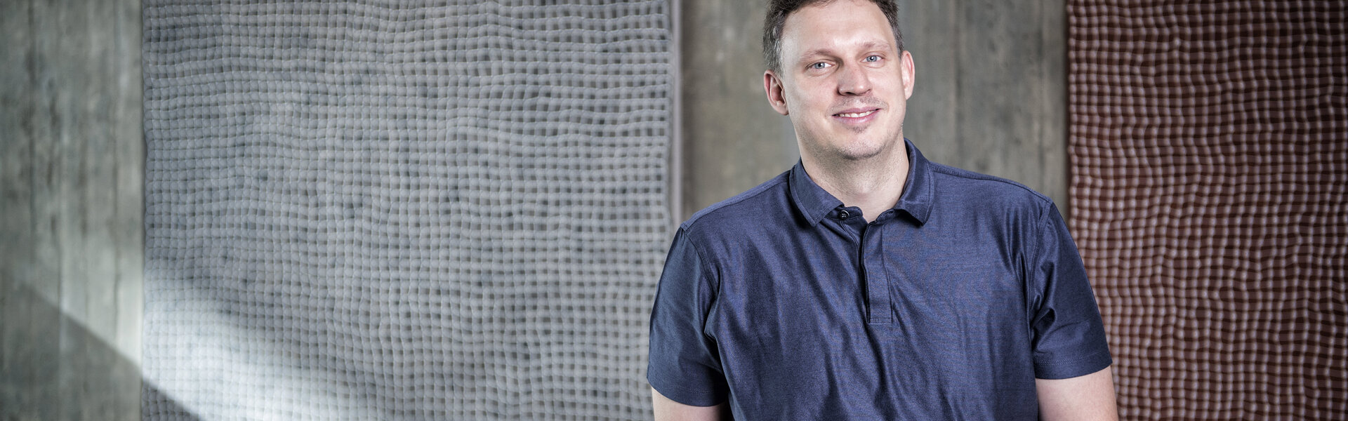 Porträtaufnahme von Ferdinand Hirschfelder in einem blauen Poloshirt vor einer Sichtbetonwand mit zwei quadratischen, strukturierten Kunstwerken.