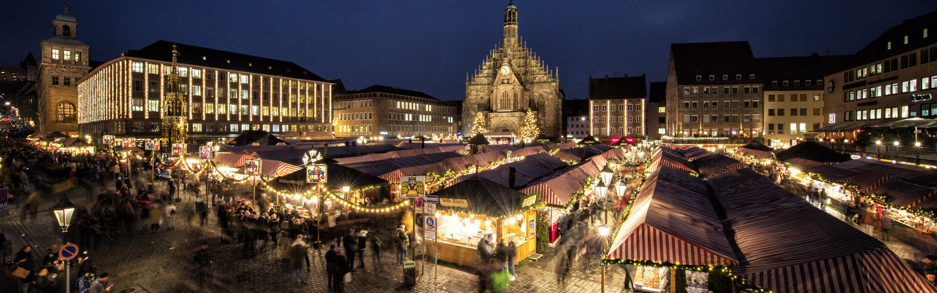 Abendlicher Blick über den Nürnberger Christkindlesmarkt mit seinen rot-weiß gestreiften Buden. Im Hintergrund erstrahlt die beleuchtete Frauenkirche vor einem tiefblauen Nachthimmel, während zahlreiche Besucher über den festlich beleuchteten Marktplatz schlendern.