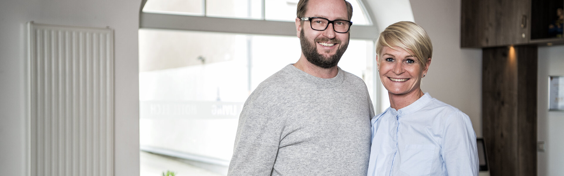 Porträtaufnahme von Robert und Eva Horka, die lächelnd in einem hellen Raum mit schwarz-weiß kariertem Fliesenboden und einem großen Rundbogenfenster im Hintergrund stehen.