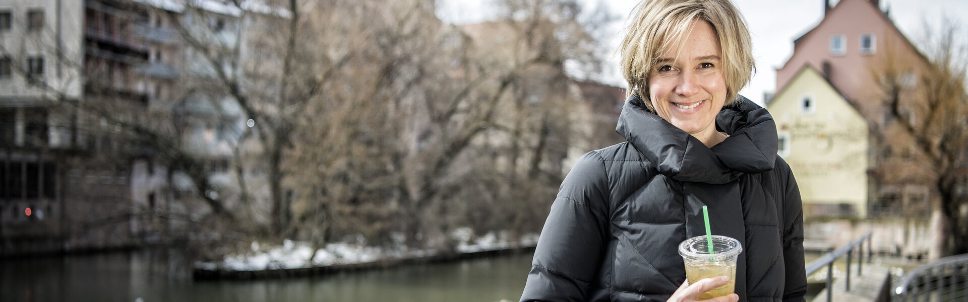 Juilia Rübsamen steht im Wintermantel an einem Gewässer und hält einen Becher mit Strohhalm, im Hintergrund verschneite Häuser und kahle Bäume.