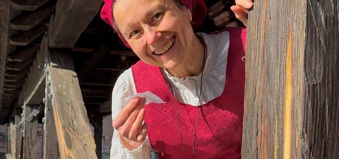 Eine als Henkersfrau Maria Schmidt verkleidete lächelnde Frau spitzt hinterm Henkersteg hervor. Sie trägt ein rotes Gewand mit weißer Bluse. Auf dem Kopf sitzt eine rote Haube.