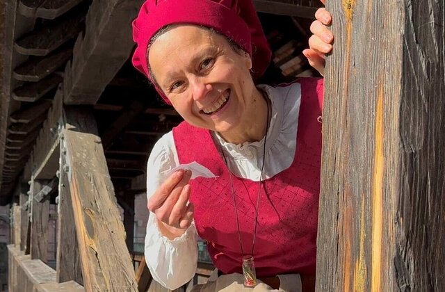 Eine als Henkersfrau Maria Schmidt verkleidete lächelnde Frau spitzt hinterm Henkersteg hervor. Sie trägt ein rotes Gewand mit weißer Bluse. Auf dem Kopf sitzt eine rote Haube.
