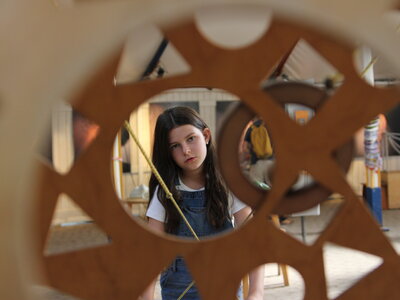 Kind mit langem Haar und Latzhose steht hinter einem großen, runden Holzbrett mit verschiedenen geometrischen Aussparungen im Erfahrungsfeld Nürnberg