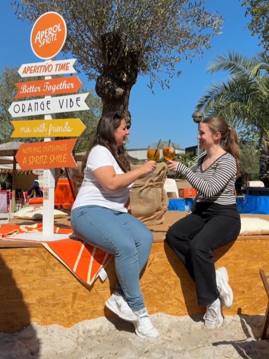 Zwei Frauen sitzen auf einer Holzbank mit orange-weißem Tuch vor einem Baum und stoßen mit Getränken an, im Hintergrund Palmen und ein bunter Wegweiser mit Aperol-Spritz-Werbung.